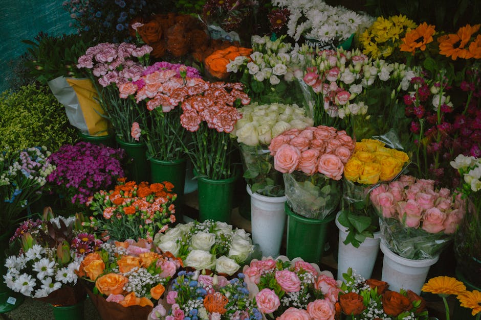 A vibrant display of fresh flowers arranged in various bouquets and potted plants, featuring roses, carnations, daisies, and other flowers in shades of pink, white, yellow, orange, and purple. The arr