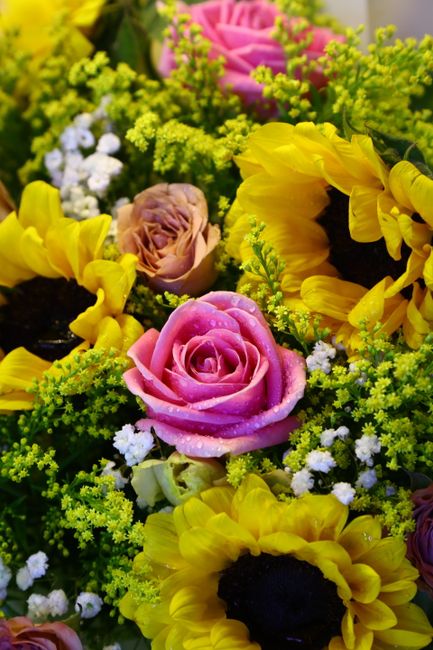 A vibrant floral arrangement featuring pink roses with dew-dropped petals, complemented by bright yellow sunflowers with dark centers, small white filler flowers, and lush green foliage. The flowers a